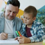 Caring father creative man is drawing pictures while kid is watching sitting at desk in house enjoying time together. Family activities and lifestyle concept.
