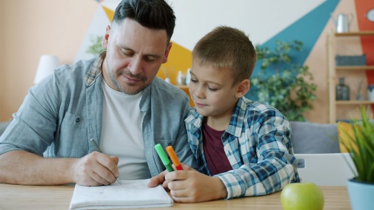 Caring father creative man is drawing pictures while kid is watching sitting at desk in house enjoying time together. Family activities and lifestyle concept.